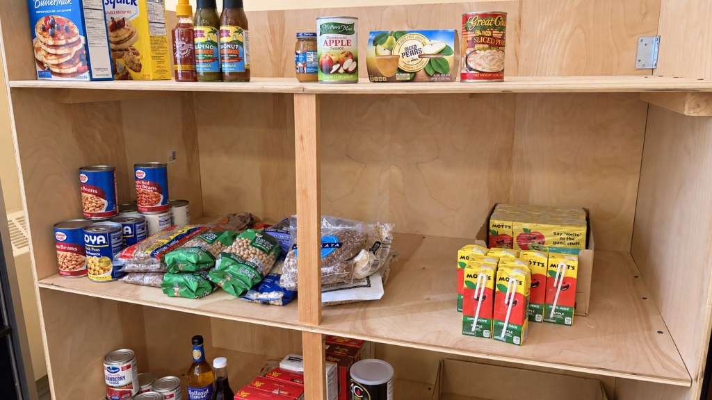 Church food pantry shelves stocked with canned and dried beans, applesauce, pancake mix, and juice boxes