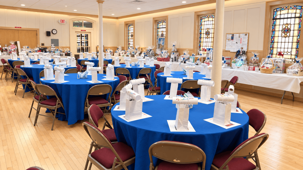 Fellowship hall set for the Holiday Tricky Tray, with blue-covered round tables, welcome gift bags, and raffle baskets under stained-glass windows