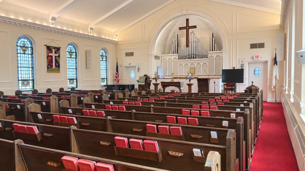 Sanctuary seen from the side, with stained-glass windows, dark wooden pews, red hymnals, and organ pipes behind the cross