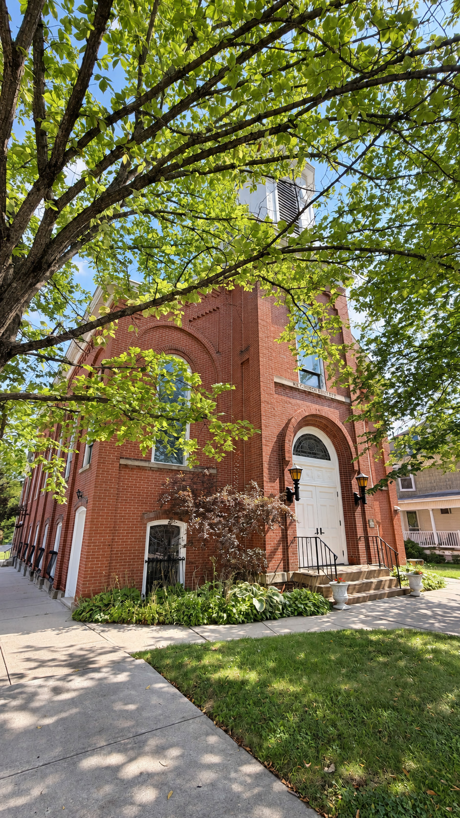 Front entrance of the red-brick church framed by green tree branches, with white double doors, lantern lights, and front steps